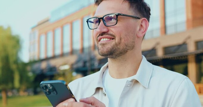 Portrait of attractive confident smiling adult dressed white shirt man holding a phone and standing outside near office building. Copy space