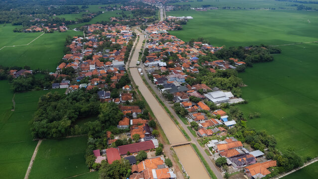 Aerial view of a village with terracotta-roofed houses nestled among vibrant green rice paddies and a central river flowing through the landscape in Indramayu, West Java, Indonesia.