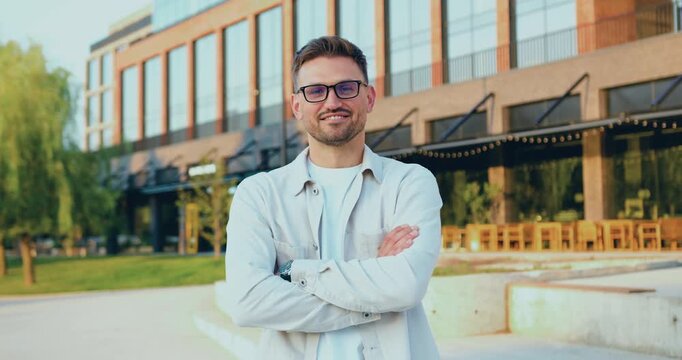 Portrait of a confident businessman in a glasses with arms crossed, looking at the camera near a modern glass office building