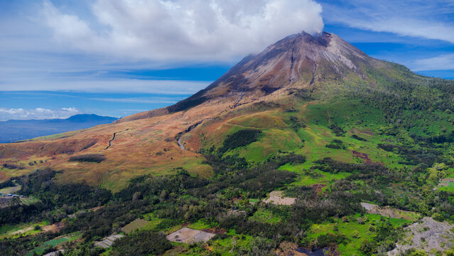 Aerial view of Mount Sinabung with volcanic smoke rising from the peak above lush green slopes and agricultural fields in Naman Teran, North Sumatra, Indonesia.