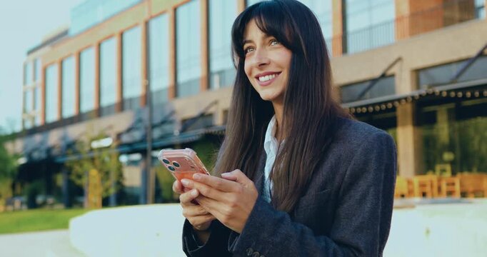 Young Caucasian businesswoman standing outside and smiling while using smartphone, with modern glass office building in the background