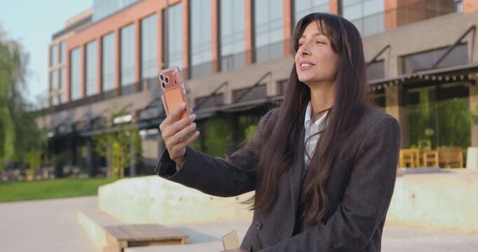 Business woman in glasses walking and having a video call on smartphone in urban business district waving hand. Female entrepreneur having an online meeting with client or business partner.