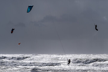 Kitesurfer springen hoch in die Luft über schäumender Brandung unter dunklem, dramatischem Wolkenhimmel an einer stürmischen Meeresküste. © IrkIngwer