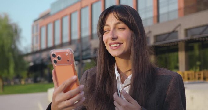 Close up of smiling Caucasian businesswoman talking on video call using phone outdoors while sitting on bench near office building. Online chat