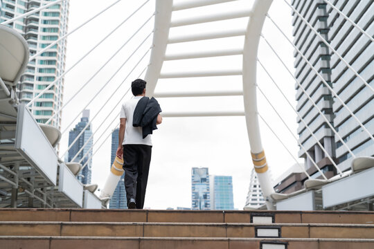 Businessman walking on modern city skywalk bridge carrying jacket over shoulder, rear view with urban skyscrapers background, confident professional commuting downtown