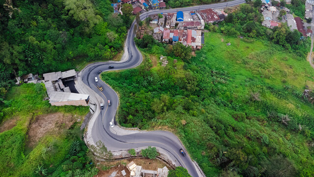 Aerial view of the winding Penatapan Berastagi road surrounded by lush green mountains, dense forest, and small buildings in Sibolangit, North Sumatra, Indonesia.
