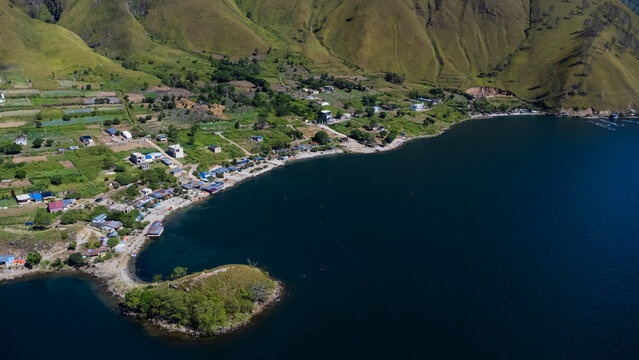 Aerial view of the scenic coastline of Lake Toba, featuring lush green hills, small village houses, and agricultural fields along the water in Paropo, Sumatera Utara, Indonesia.