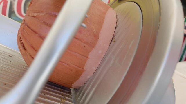 Close-up of a cured meat roll held by metal prongs on a deli slicer