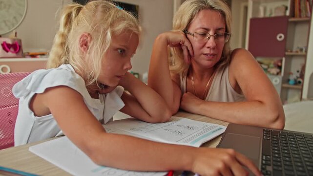 Schoolgirl using laptop for homework with mother sitting nearby, workbook open on desk. Remote learning and home education