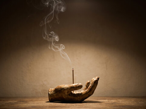 Ceramic hand incense burner with burning incense stick and delicate rising smoke on rustic wooden table against warm dark background