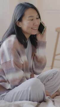 Cheerful Asian woman speaking on mobile phone laughing sitting on bed in luxurious apartment. Cellphone communication and people concept.
