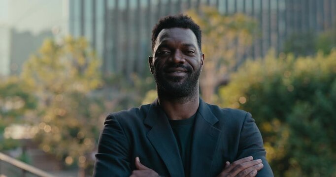 Confident African American businessman stands with crossed arms outside a modern office, looking at the camera in a strong corporate leadership portrait setting.