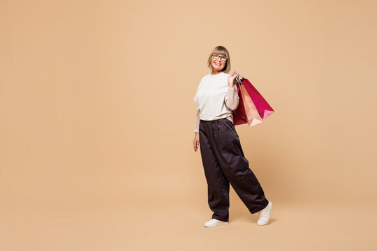 Full body elderly happy young woman wear casual clothes hold in hand paper package bags after shopping look aside isolated on plain pastel light beige background. Black Friday sale buy day concept