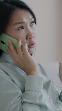 Young Asian woman making mobile phone call sitting on couch in luxury apartment. Cellphone communication and wireless technology concept.
