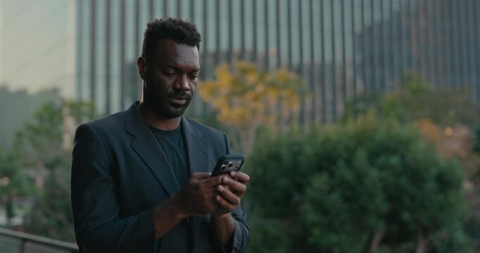 Smiling African American businessman types a message on his smartphone outside a modern office, enjoying convenient mobile communication during the workday in the city.