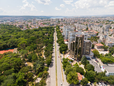 Aerial view of a wide boulevard lined with lush green trees in the Sea Garden park, adjacent to modern high-rise buildings and the urban cityscape of Varna, Varna, Bulgaria.