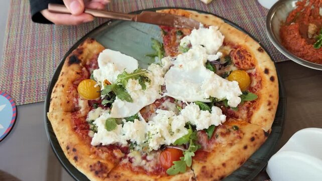 Close-up top-down view of a hand holding a knife and cutting a freshly baked burrata pizza topped with tomatoes and arugula on a restaurant dining table during daytime.