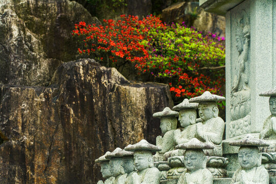 Rows of small stone Buddhist monk statues in prayer beside a rocky cliff with blooming azaleas in a traditional temple.