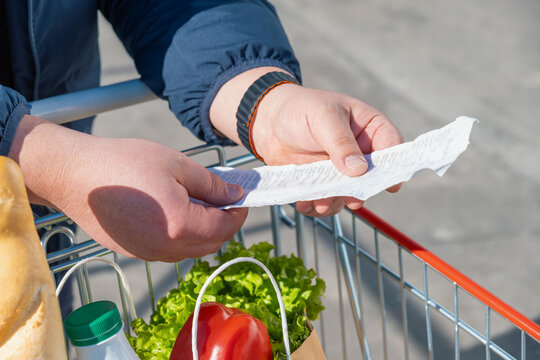 Shopper holding and checking long supermarket receipt above cart with groceries. Everyday food purchases, rising grocery prices, inflation, increasing cost of living and growing consumer budget