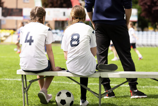 Youth Girls Soccer Substitutes in White Jerseys 4 and 8 Sitting on Sideline Bench with Coach Standing Beside Them - Junior Football Team