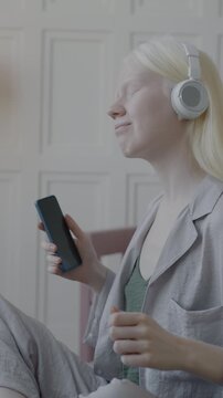 Slow motion of happy albino girl in pajamas listening to music through headphones and dancing holding smartphone in bed in luxury bedroom. Youth culture and leisure fun concept.