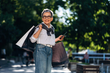 Obraz premium Senior Asian woman carrying shopping bags and holding a smartphone, smiling while standing outdoors in a park with green trees and blurred background