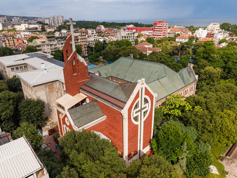 Aerial view of the Evangelical Methodist Episcopal Church with its distinctive red brick architecture and cross, surrounded by lush green trees in the urban landscape of Varna, Varna, Bulgaria.
