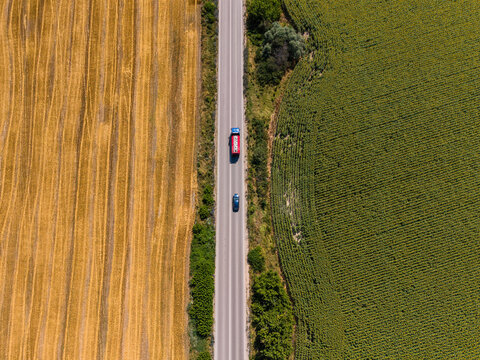 Aerial view of a road running between a golden harvested field and a green field with a red truck and a blue car driving in Devnya, Varna, Bulgaria.