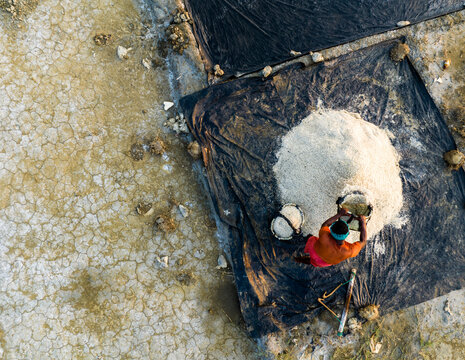 Aerial view of a salt worker collecting salt from a large pile on a black plastic sheet in a salt field in Moheskhali Upazila, Chittagong Division, Bangladesh.