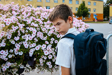 Young boy with backpack stands beside blooming pink flowers in front of a school building, looking away from the camera on a sunny day © Jolanta