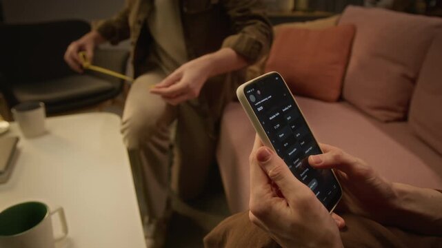 Cropped shot of hands of unrecognizable woman browsing furniture catalog on smartphone while partner measures room with tape measure. For mobile retail advertising and interior design app promotion