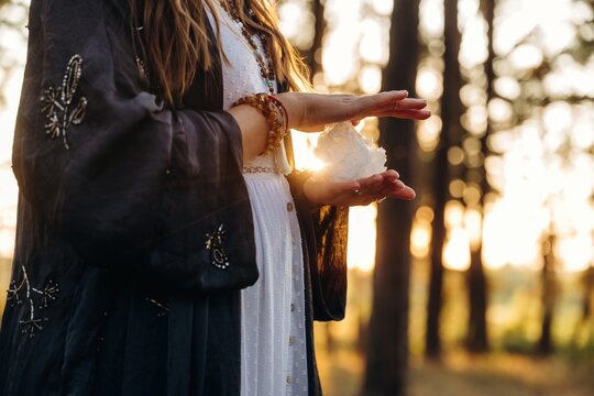 Woman in flowing white dress and dark shawl holds a crystal in her hands, surrounded by tall trees and soft sunlight during golden hour in a natural setting