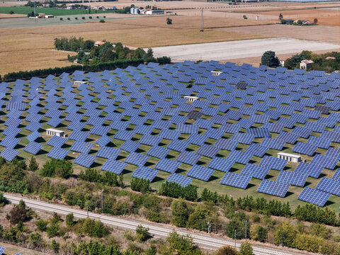 Aerial view of a large solar panel system in a rural field with rows of photovoltaic panels, surrounding agricultural land, and a nearby railway line in Spinetta Marengo, Piedmont, Italy.