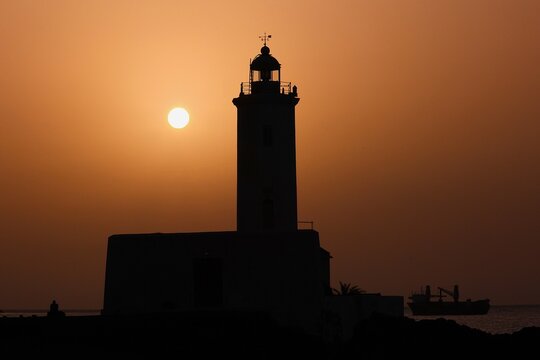 Silhouette d'un phare isol&eacute; en bordure de mer avec le soleil en contre jour. Clich&eacute; pris au l'aube avec un ciel orang&eacute; en arri&egrave;re plan.