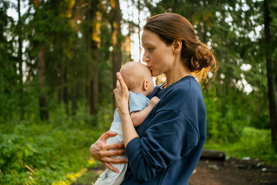 Mother tenderly holding her baby son and giving a kiss on his forehead, embracing maternal love and connection outdoors in a green forest bathed in golden light