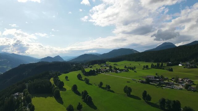 Aerial drone view of the idyllic countryside near Ramsau am Dachstein in Austria. Slow flight over lush green alpine meadows, traditional mountain houses and forests illuminated by warm summer evening