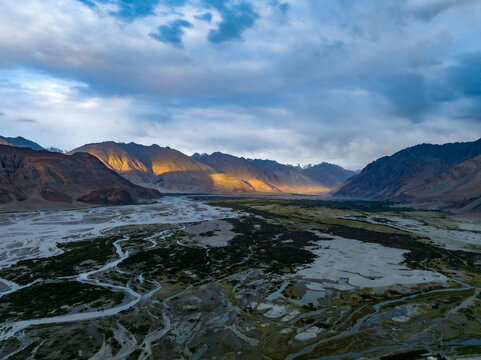 Aerial view of the rugged mountain landscape of Ladakh with a winding riverbed and dramatic golden light hitting the peaks under a cloudy sky in Ladakh Division, Ladakh, India.
