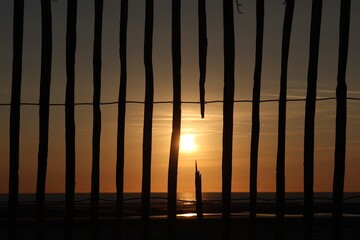 Vue rapprochée de la silhouette d'une palissade en bois avec une ouverture laissant passer le soleil couchant. Forme géométrique avec des éléments verticaux. © Xavier