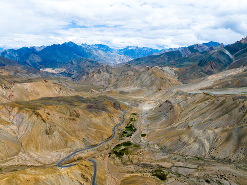 Aerial view of the rugged Himalayan mountain range featuring winding roads through arid valleys, rocky terrain, and distant snow-capped peaks in Ladakh, India.