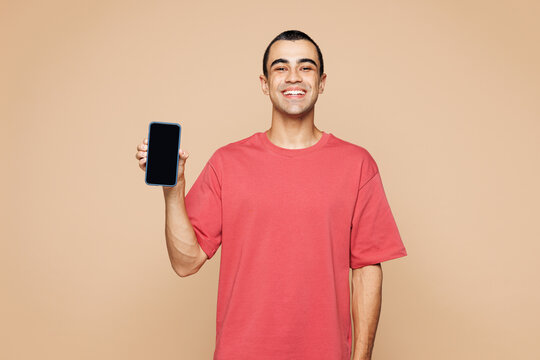 Young middle eastern man wear red t-shirt casual clothes hold in hand use mobile cell phone with blank screen workspace area isolated on plain pastel light beige background studio. Lifestyle concept