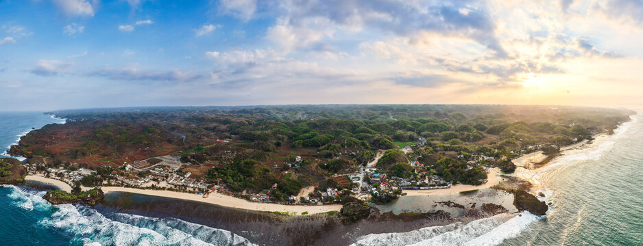 Aerial view of the rugged coastline with lush green hills, sandy beach, and crashing waves under a golden sunset in Gunungkidul Regency, Special Region of Yogyakarta, Indonesia.