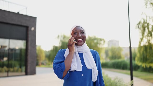A happy African American Muslim woman wearing a hijab and blue dress talks on her smartphone outdoors