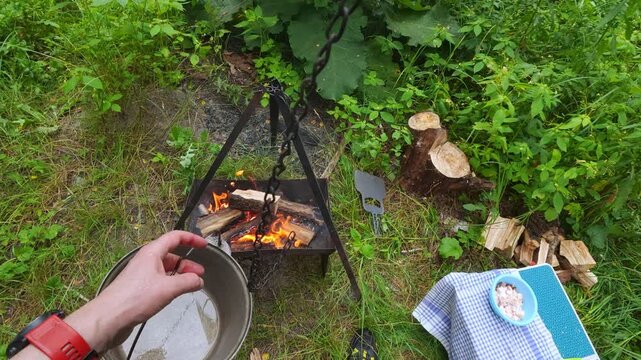 Campers hanging metal cauldron on tripod chain over campfire for outdoor cooking