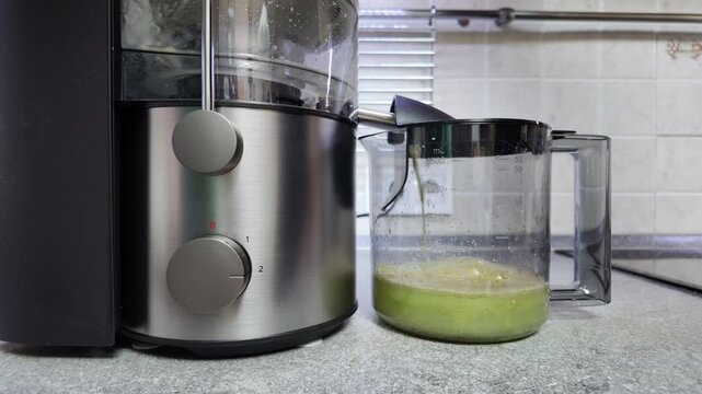 Electric juicer extracting fresh green juice filling transparent pitcher in kitchen, time-lapse video