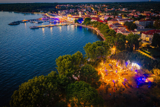 Aerial view of the illuminated harbor and town of Fazana at night, featuring docked boats, historic buildings, and an outdoor event with string lights. Fazana, Istria County, Croatia.