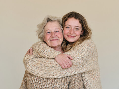 Happy young granddaughter hugging her elderly grandmother tightly from behind against a neutral light background