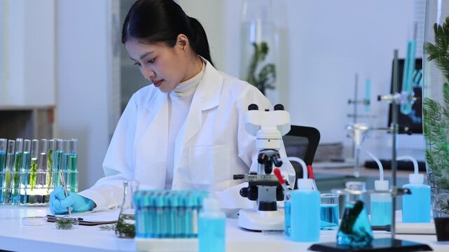 Focused asian female botanist writing research data in a modern science laboratory with a microscope, test tubes, plants, and colorful liquid for biotechnology development