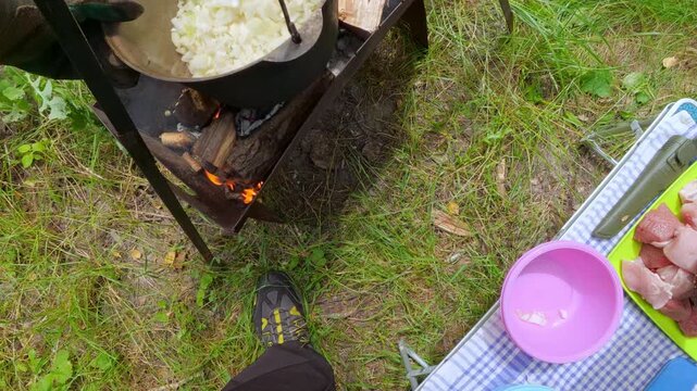Man adding chopped onion into campfire cauldron for outdoor picnic cooking lifestyle