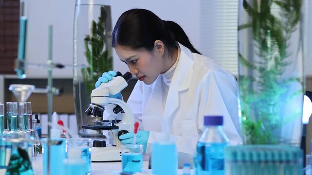 Young female biologist researcher looking into a microscope in a laboratory, developing a new cosmetic product from algae and other plants with glassware and test tube