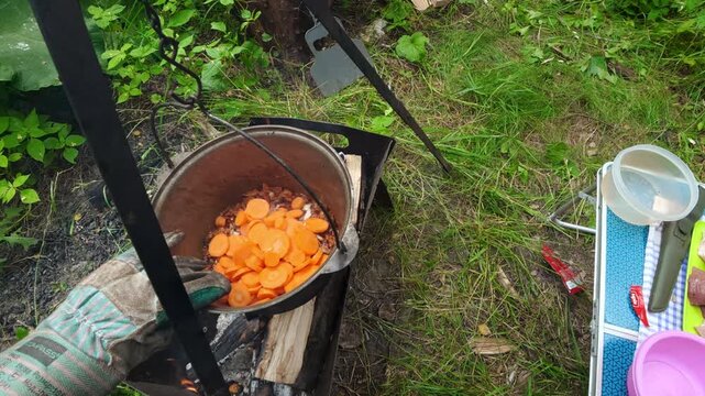 Man adding sliced fresh carrots into cauldron over campfire for outdoor camping cooking lifestyle